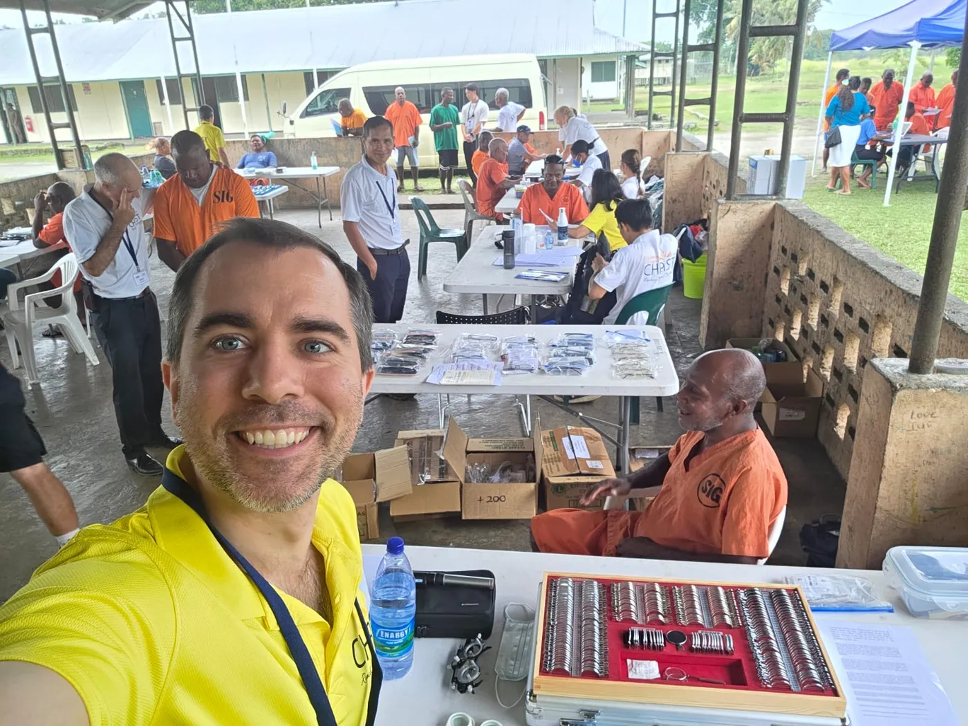 Tom at an outdoor eye clinic, with a case of trial lenses and patients waiting behind him