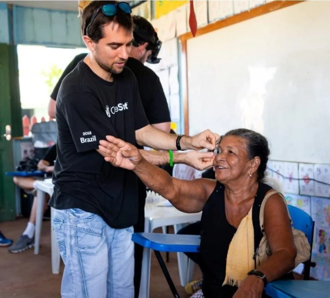 Tom guiding an older woman through a vision check at a clinic table